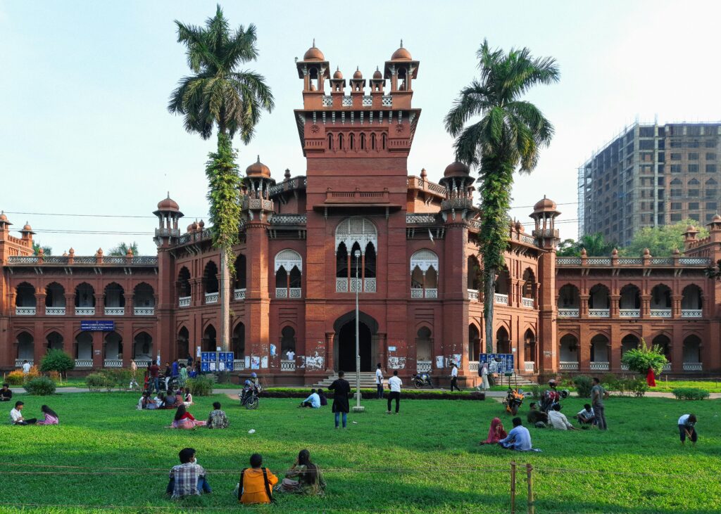 Historic red-brick architecture at the University of Dhaka with people on the lawn enjoying a sunny day.